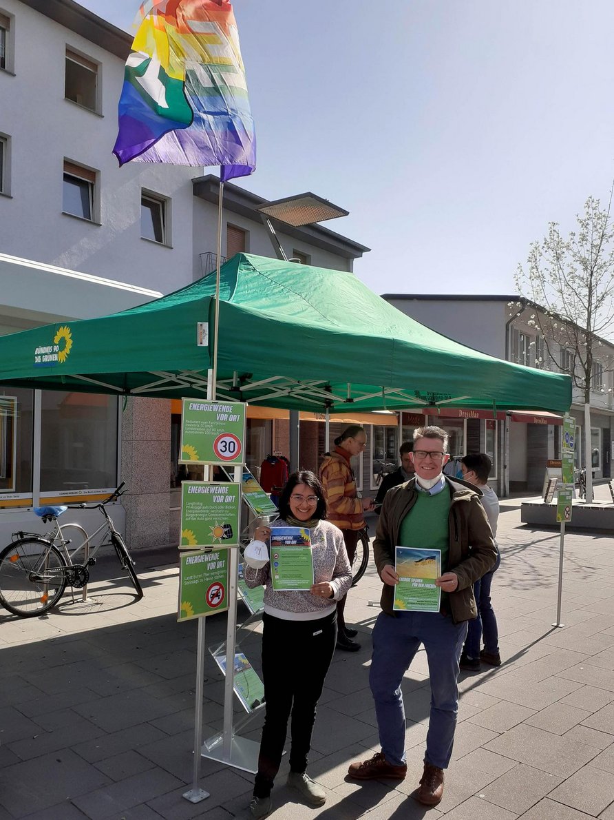 Infostand von Bündnis 90/Die Grünen. Neben einem Prospektständer stehen Melanie Middendorf Pelaiz (Sprecherin OV Heidesheim) und Jörn Siemon (Pressesprecher OV Ingelheim). Beide halten den Flyer zum Infostand "Energiesparen für den Frieden" in der Hand. Im Hintergrund steht Heinrich Jung (Fraktionsvorsitzender) und ist im Gespräch mit einem Passanten. Im Bild oben angeschnitten ist eine Regenbogenfahne mit der Aufschrift Peace.